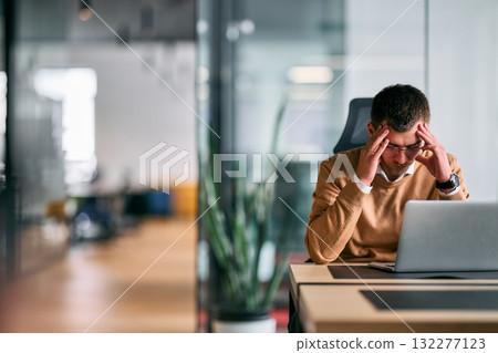 Concentrated businessman sitting at desk using laptop in modern office interior, thinking and analyzing data or business strategy, symbolizing productivity, focus, and professional work. 132277123