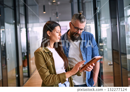 Two business professionals having a focused discussion while reviewing information on a tablet in a bright modern office hallway, symbolizing teamwork, communication, and digital collaboration. 132277124