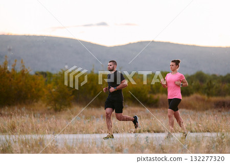 A couple dressed in sportswear runs along a scenic road during an early morning workout, enjoying the fresh air and maintaining a healthy lifestyle 132277320