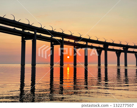 Beautiful sunset over the sea. The sun setting between the pillars of the bridge. Reflection of sunlight on water. The sky is painted in golden colors. Kanonersky Island, Saint-Petersburg Beautiful sunset over the sea. The sun setting between the pillars of the bridge. Reflection of sunlight on water. The sky is painted in golden colors. Kanonersky Island, Saint-Petersburg 132277507