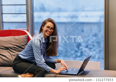 A businesswoman utilizes her laptop while seated by the window of a large corporate building, offering a picturesque view of the city skyline as her backdrop. 132277853