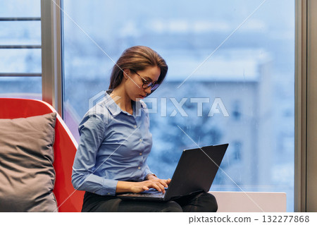 A businesswoman utilizes her laptop while seated by the window of a large corporate building, offering a picturesque view of the city skyline as her backdrop. 132277868