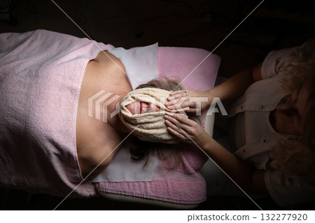 Close up of a mature woman receiving a relaxing facial massage from a professional female therapist in a modern spa. The client lies on a treatment bed with a towel and headband, enjoying skincare Close up of a mature woman receiving a relaxing facial massage from a professional female therapist in a modern spa. The client lies on a treatment bed with a towel and headband, enjoying skincare 132277920
