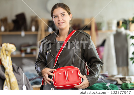 Winter collection sale. Positive young girl client standing against background of shelves with clothes in modern store and choosing red bag Winter collection sale. Positive young girl client standing against background of shelves with clothes in modern store and choosing red bag 132277947