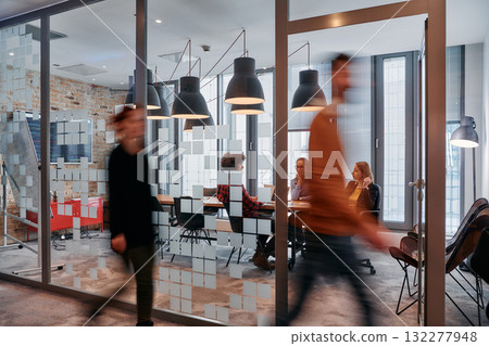 In the dynamic hustle and bustle of a business environment, a group of young business professionals walk down the corridor next to their office, while their colleagues collaborate inside, symbolizing 132277948