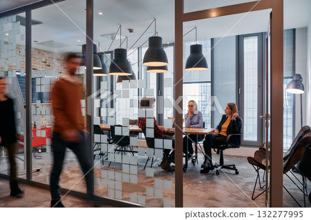 In the dynamic hustle and bustle of a business environment, a group of young business professionals walk down the corridor next to their office, while their colleagues collaborate inside, symbolizing 132277995