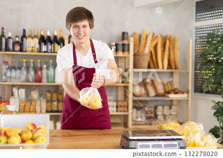In store, guy worker in striped apron takes apples from drawer and puts them in disposable bag. In store, guy worker in striped apron takes apples from drawer and puts them in disposable bag. 132278020