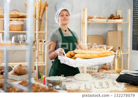 Bakery female seller stands proudly in front of fresh bread holding basket of freshly baked baguettes in kitchen Bakery female seller stands proudly in front of fresh bread holding basket of freshly baked baguettes in kitchen 132278079