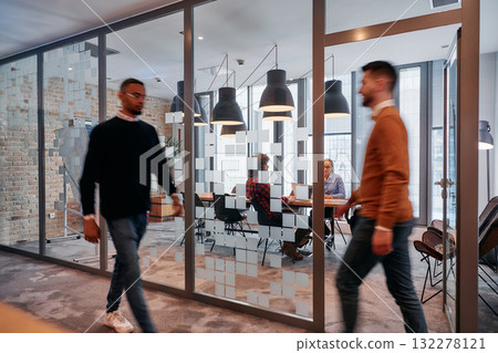 In the dynamic hustle and bustle of a business environment, a group of young business professionals walk down the corridor next to their office, while their colleagues collaborate inside, symbolizing 132278121