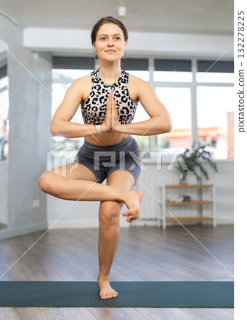Young girl doing hatha yoga in modern studio, performing balancing asana Vrikshasana with hands clasped in namaste 132278225