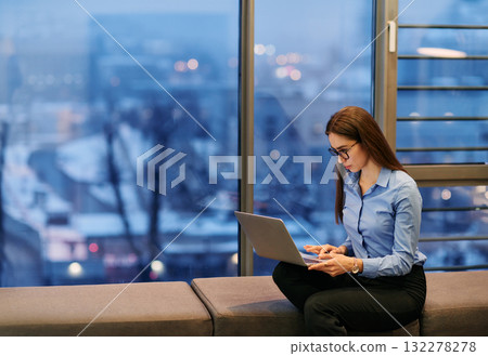A businesswoman utilizes her laptop while seated by the window of a large corporate building, offering a picturesque view of the city skyline as her backdrop. 132278278
