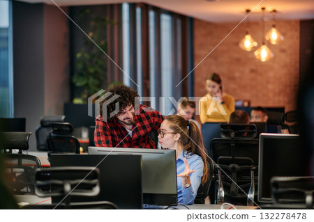 Business colleagues, a man and a woman, engage in discussing business strategies while attentively gazing at a computer monitor, epitomizing collaboration and innovation Business colleagues, a man and a woman, engage in discussing business strategies while attentively gazing at a computer monitor, epitomizing collaboration and innovation 132278478