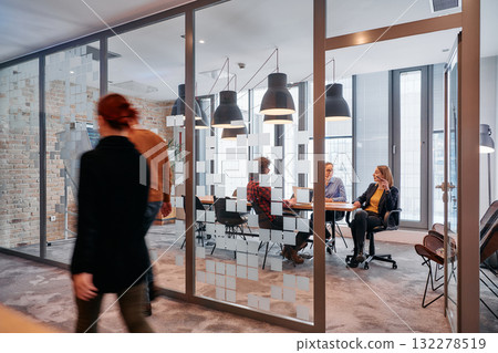 In the dynamic hustle and bustle of a business environment, a group of young business professionals walk down the corridor next to their office, while their colleagues collaborate inside, symbolizing 132278519