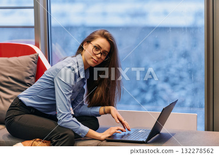 A businesswoman utilizes her laptop while seated by the window of a large corporate building, offering a picturesque view of the city skyline as her backdrop. 132278562