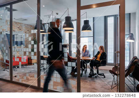 In the dynamic hustle and bustle of a business environment, a group of young business professionals walk down the corridor next to their office, while their colleagues collaborate inside, symbolizing 132278648