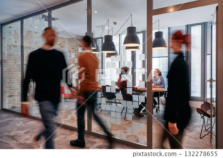 In the dynamic hustle and bustle of a business environment, a group of young business professionals walk down the corridor next to their office, while their colleagues collaborate inside, symbolizing 132278655