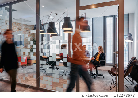 In the dynamic hustle and bustle of a business environment, a group of young business professionals walk down the corridor next to their office, while their colleagues collaborate inside, symbolizing 132278678