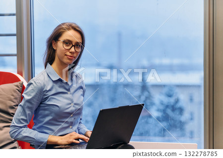 A businesswoman utilizes her laptop while seated by the window of a large corporate building, offering a picturesque view of the city skyline as her backdrop. A businesswoman utilizes her laptop while seated by the window of a large corporate building, offering a picturesque view of the city skyline as her backdrop. 132278785