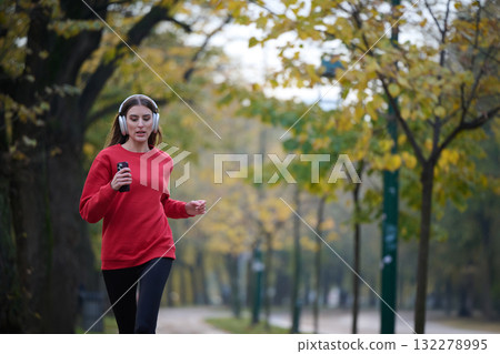 Young beautiful woman running in autumn park and listening to music with headphones on smartphone 132278995
