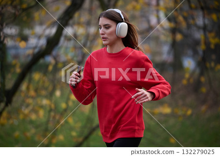 Young beautiful woman running in autumn park and listening to music with headphones on smartphone 132279025