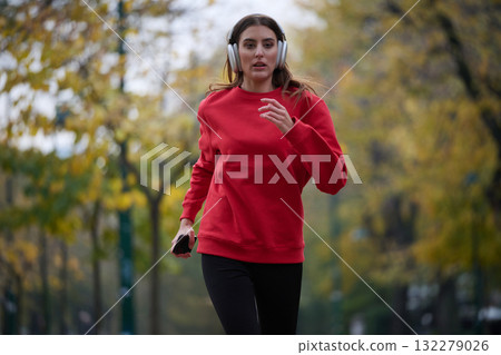 Young beautiful woman running in autumn park and listening to music with headphones on smartphone 132279026