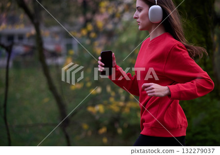 Young beautiful woman running in autumn park and listening to music with headphones on smartphone 132279037