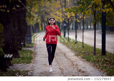 Young beautiful woman running in autumn park and listening to music with headphones on smartphone 132279047