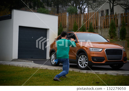 A man meticulously washes and cleans his car in the driveway of his home, showcasing pride in his vehicle and outdoor space. 132279050