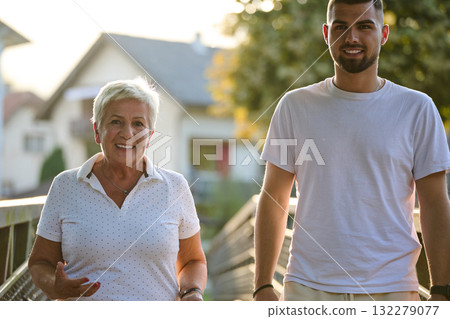 A handsome man and an older woman share a serene walk in nature, crossing a beautiful bridge against the backdrop of a stunning sunset, embodying the concept of a healthy and vibrant intergenerational 132279077