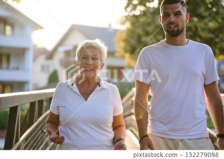 A handsome man and an older woman share a serene walk in nature, crossing a beautiful bridge against the backdrop of a stunning sunset, embodying the concept of a healthy and vibrant intergenerational A handsome man and an older woman share a serene walk in nature, crossing a beautiful bridge against the backdrop of a stunning sunset, embodying the concept of a healthy and vibrant intergenerational 132279098