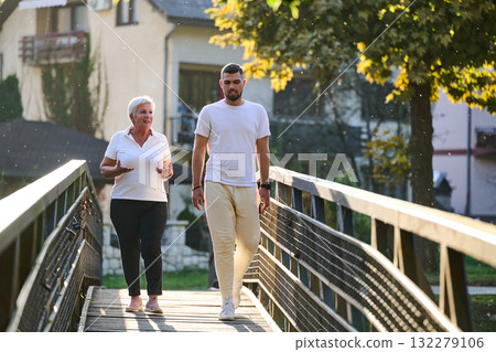 A handsome man and an older woman share a serene walk in nature, crossing a beautiful bridge against the backdrop of a stunning sunset, embodying the concept of a healthy and vibrant intergenerational A handsome man and an older woman share a serene walk in nature, crossing a beautiful bridge against the backdrop of a stunning sunset, embodying the concept of a healthy and vibrant intergenerational 132279106