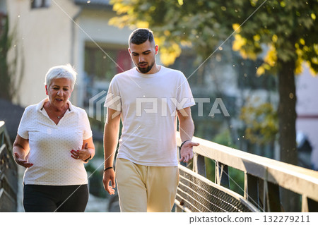A handsome man and an older woman share a serene walk in nature, crossing a beautiful bridge against the backdrop of a stunning sunset, embodying the concept of a healthy and vibrant intergenerational 132279211