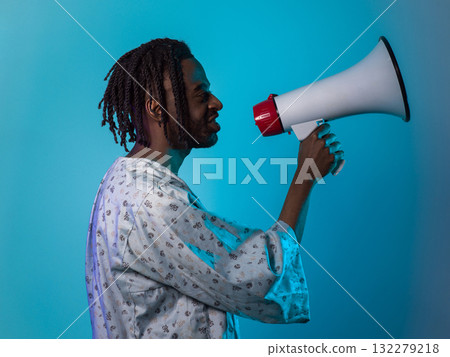 African American man dons traditional attire, passionately utilizing a megaphone against a striking blue background, symbolizing his vocal and cultural empowerment in the pursuit of social justice and 132279218