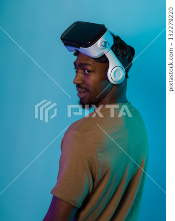 In a futuristic visual, an African American man stands isolated against a striking blue backdrop, adorned with VR glasses that transport him into a cutting-edge virtual reality experience, merging 132279220
