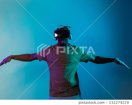 African American man dons VR glasses, engrossed in a virtual reality simulation, gesturing with hands, against a striking blue background, highlighting the fusion of cutting-edge technology and African American man dons VR glasses, engrossed in a virtual reality simulation, gesturing with hands, against a striking blue background, highlighting the fusion of cutting-edge technology and 132279229
