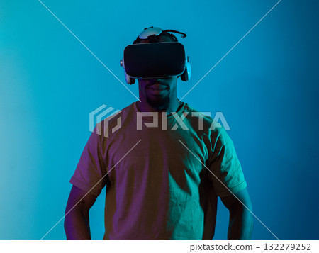 In a futuristic visual, an African American man stands isolated against a striking blue backdrop, adorned with VR glasses that transport him into a cutting-edge virtual reality experience, merging In a futuristic visual, an African American man stands isolated against a striking blue backdrop, adorned with VR glasses that transport him into a cutting-edge virtual reality experience, merging 132279252