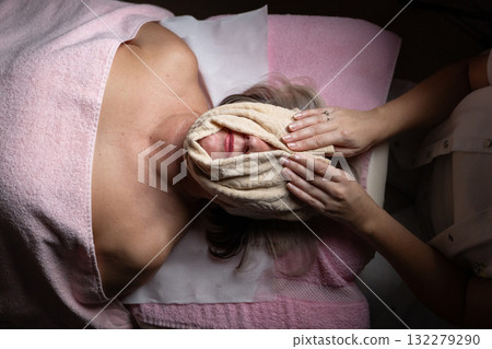 Close up of a mature woman receiving a relaxing facial massage from a professional female therapist in a modern spa. The client lies on a treatment bed with a towel and headband, enjoying skincare 132279290