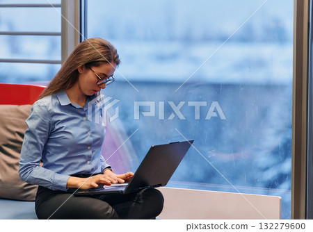 A businesswoman utilizes her laptop while seated by the window of a large corporate building, offering a picturesque view of the city skyline as her backdrop. 132279600