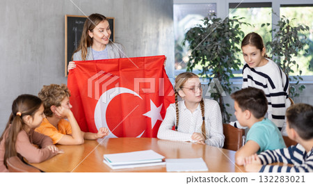 Preteens pupils looking at national flag of Turkey while learning different cultures with positive young female teacher in schoolroom 132283021
