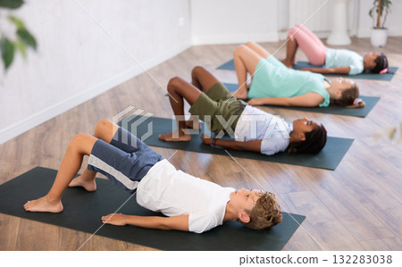 In sport studio with group of friends, boy perform bridge pose, setu bandha sarvangasana In sport studio with group of friends, boy perform bridge pose, setu bandha sarvangasana 132283038