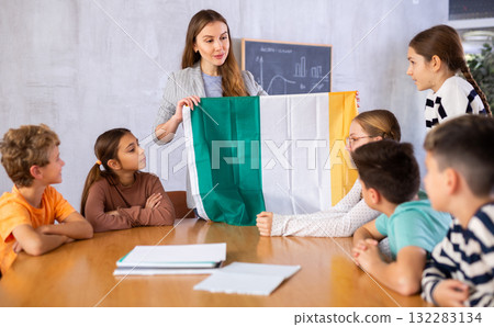 Positive young woman teacher showing national flag of Ireland and telling preteens schoolchildren history of country during lesson in class Positive young woman teacher showing national flag of Ireland and telling preteens schoolchildren history of country during lesson in class 132283134