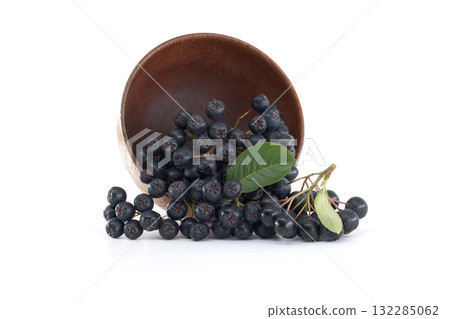 A group of aronia berries spills from a wooden bowl, isolated against a white backdrop. 132285062