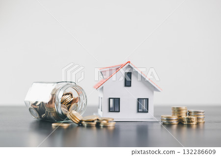 Glass jar with spilled coins beside house model, symbolizing savings, mortgage loan, property finance, real estate investment, home ownership, wealth, money management concept on minimal background. Glass jar with spilled coins beside house model, symbolizing savings, mortgage loan, property finance, real estate investment, home ownership, wealth, money management concept on minimal background. 132286609