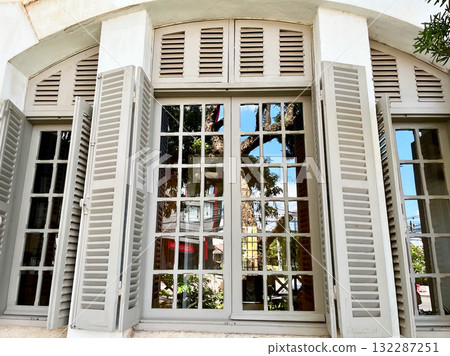Vintage French-style window with open wooden shutters and glass panes reflecting blue sky and trees outside. Architectural detail of old colonial building showing charm and natural light Vintage French-style window with open wooden shutters and glass panes reflecting blue sky and trees outside. Architectural detail of old colonial building showing charm and natural light 132287251