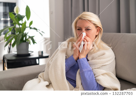 Ill woman covered with a warm plaid sitting on sofa and blowing her nose with tissue. Concept of flu, cold, home treatment, and seasonal illness during epidemic. Ill woman covered with a warm plaid sitting on sofa and blowing her nose with tissue. Concept of flu, cold, home treatment, and seasonal illness during epidemic. 132287703