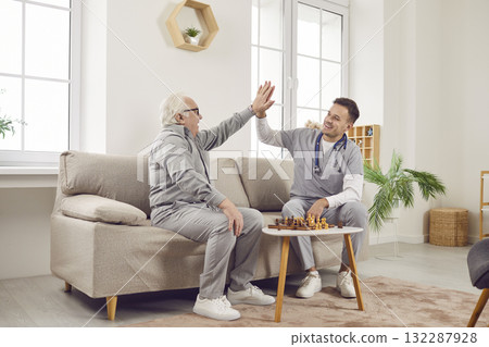 Nurse man giving high five to a gray-haired senior elderly person while playing chess game at home. 132287928