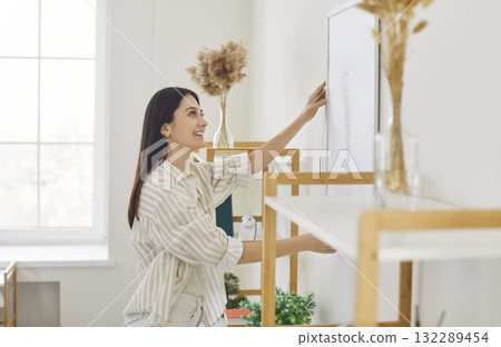 Young woman hanging picture frame on white wall, decorating her modern living space 132289454
