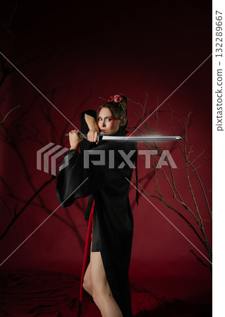 Woman poses in studio wearing traditional Japanese dress with a sword and striking a strong stance Woman poses in studio wearing traditional Japanese dress with a sword and striking a strong stance 132289667