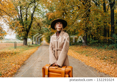 European woman in coat and hat walks along  yellow autumn trees 132291024