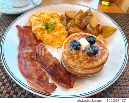 American breakfast with scrambled eggs, bacon, pancakes with blueberries, and roasted potatoes. Morning meal concept. 132292487